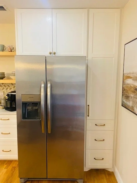 Stainless steel refrigerator in a white kitchen with upper and lower cabinets, a countertop with kitchen items, and a wall painting.