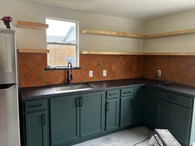 Kitchen with dark green cabinets, a black countertop, tiled backsplash, a window above the sink, and floating wooden shelves on the wall.