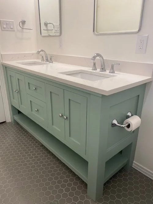 Light green double vanity bathroom with white countertop and two sinks with silver faucets, two mirrors above, a toilet paper roll holder on the side, hexagonal tile flooring.