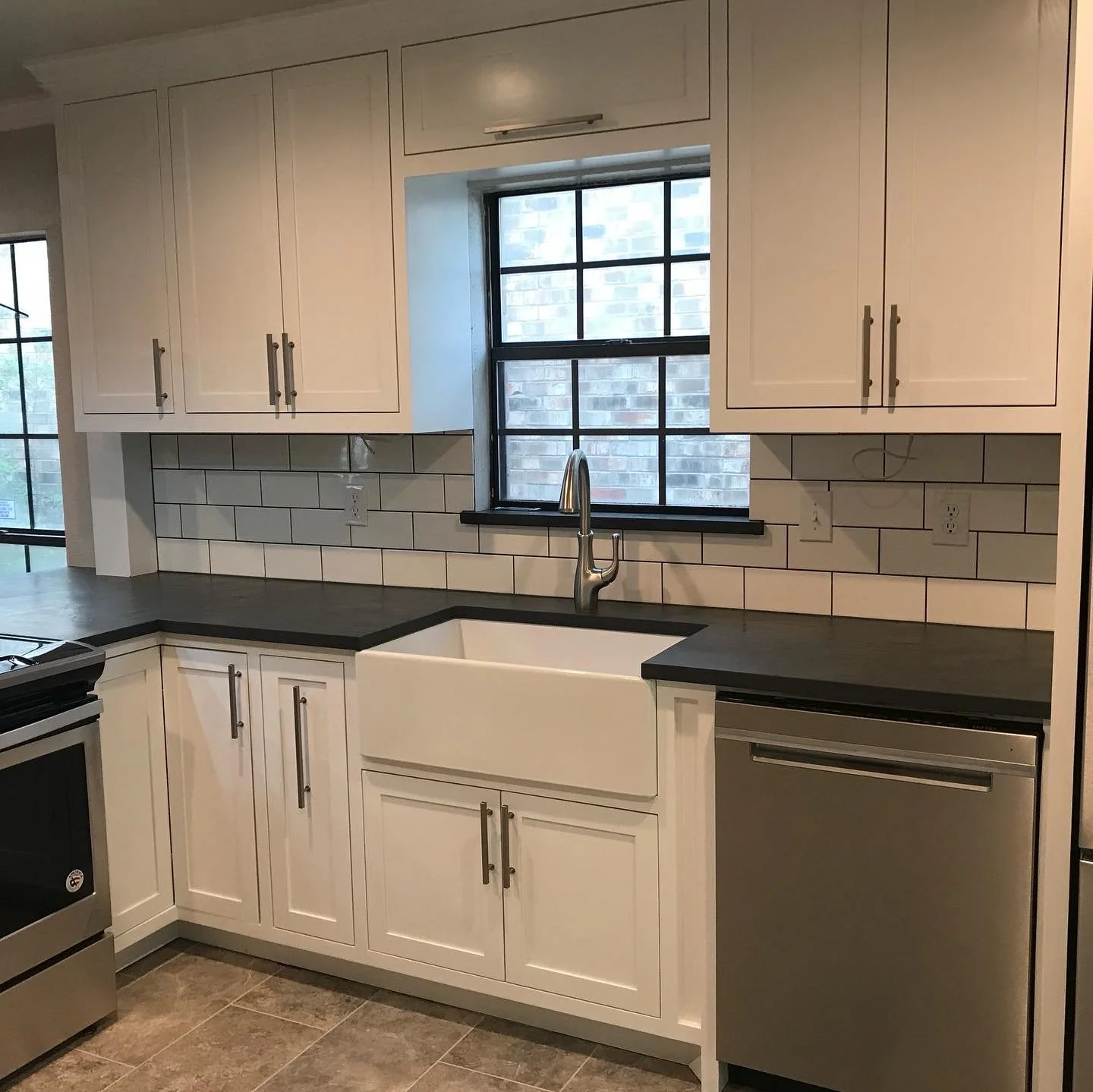 Kitchen with white cabinets, black countertops, a farmhouse sink, a window above the sink, a stainless steel dishwasher, and a brick wall visible through the window.