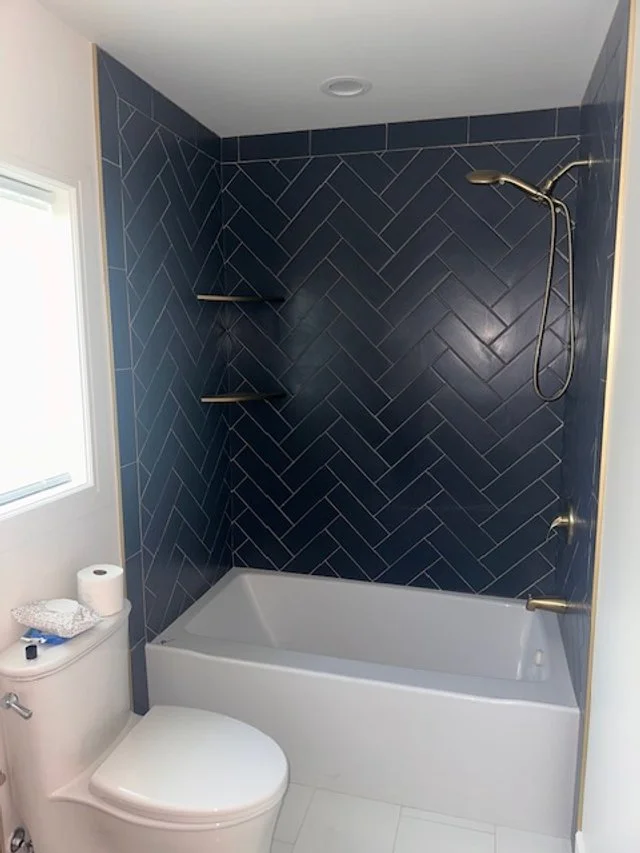 Bathroom with a white bathtub, navy blue tiled wall with diagonal pattern, a showerhead, a window, a toilet, and shelves.