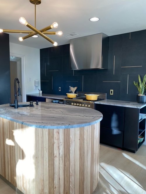 Modern kitchen with black cabinets, a marble island with a wooden facade, stainless steel range hood, and a gold chandelier. There are two yellow pots on the stove, a potted plant on the counter, and sunlight streaming in.