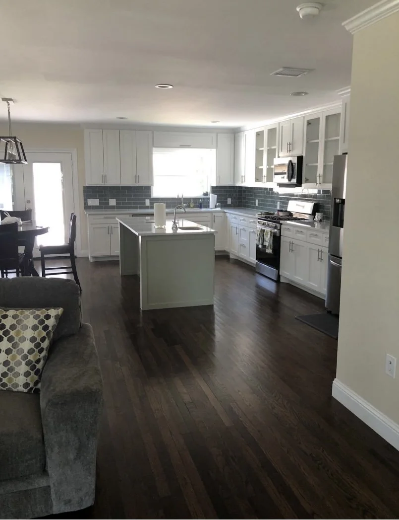Open kitchen with white cabinets, dark gray backsplash, stainless steel appliances, and dark hardwood floors. There is an island with a sink, a window above the sink, and a dining area with a table and chairs.