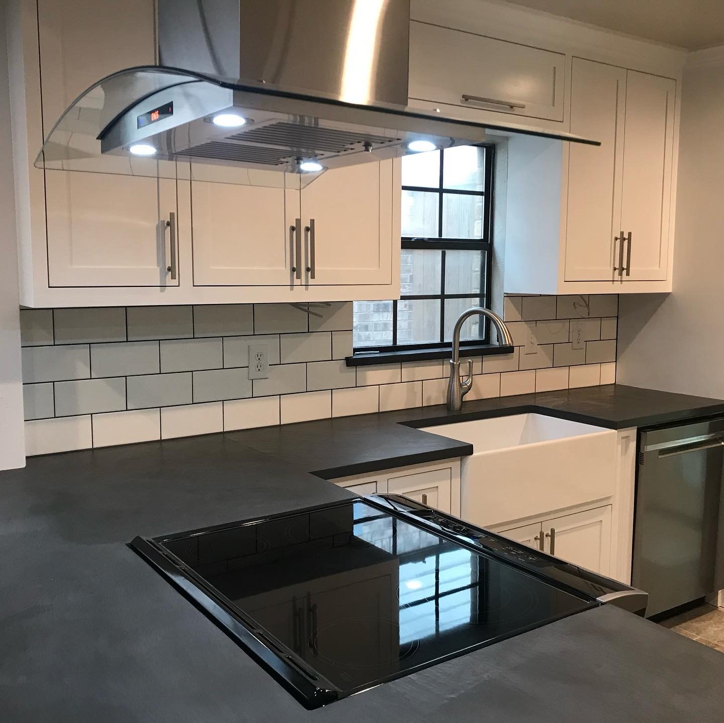 Modern kitchen with white cabinets, black countertops, a white farmhouse sink, black window, and stainless steel appliances, including a stove and dishwasher.