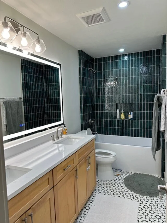 Bathroom with wooden vanity, large mirror, black tiled shower area, white bathtub, toilet, towel rack, gray towels, gray round bath rug, and patterned black and white floor tiles.
