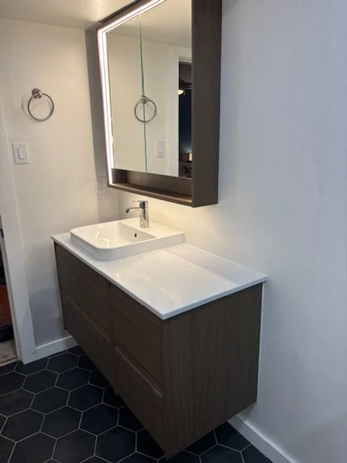 Modern bathroom sink vanity with a white countertop, vessel sink, and a large illuminated mirror; dark wood cabinet, hexagon black tile floor, and a towel ring on the wall.