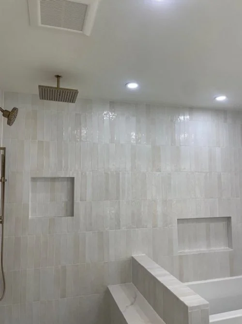 A modern walk-in shower with beige tiled walls, a square rain showerhead, a wall-mounted handheld shower, two built-in inset shelves, and a partial bench.