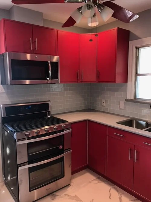 Red kitchen cabinets, stainless steel microwave and oven, white subway tile backsplash, gray countertop, and a window above the sink.
