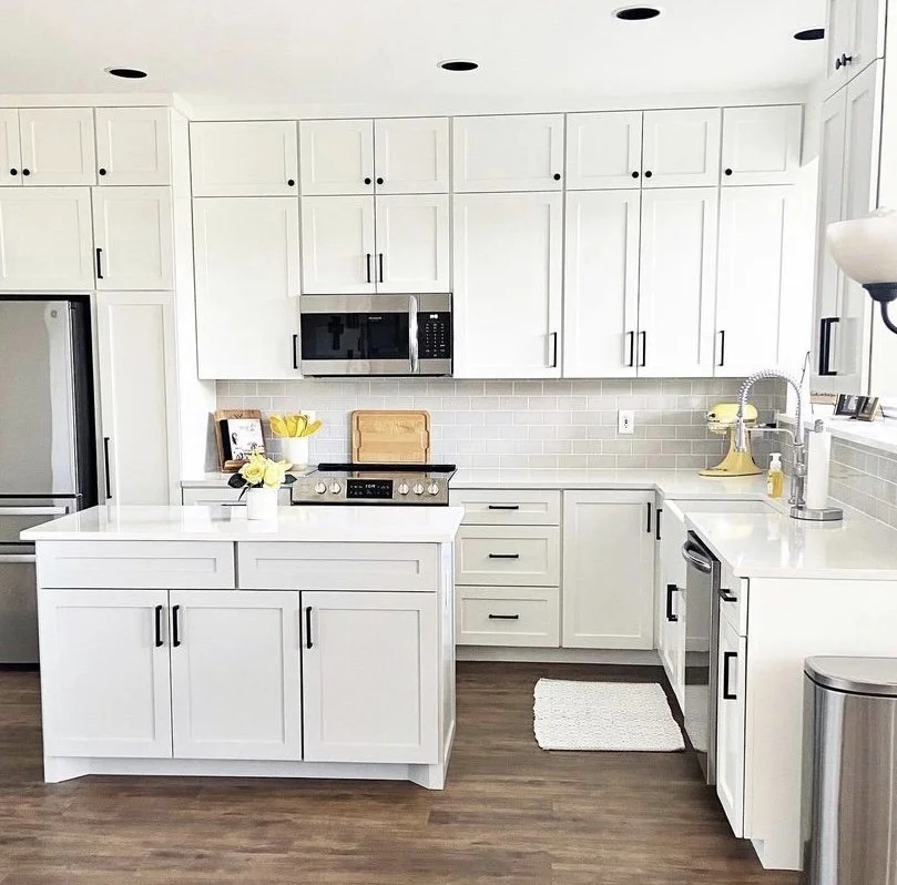 Modern kitchen with white cabinets, a white countertop, stainless steel appliances, and a wood-look floor. Kitchen items include a microwave, a stove, and a sink with a yellow dish drying rack. There is a small island with a flower in a vase.
