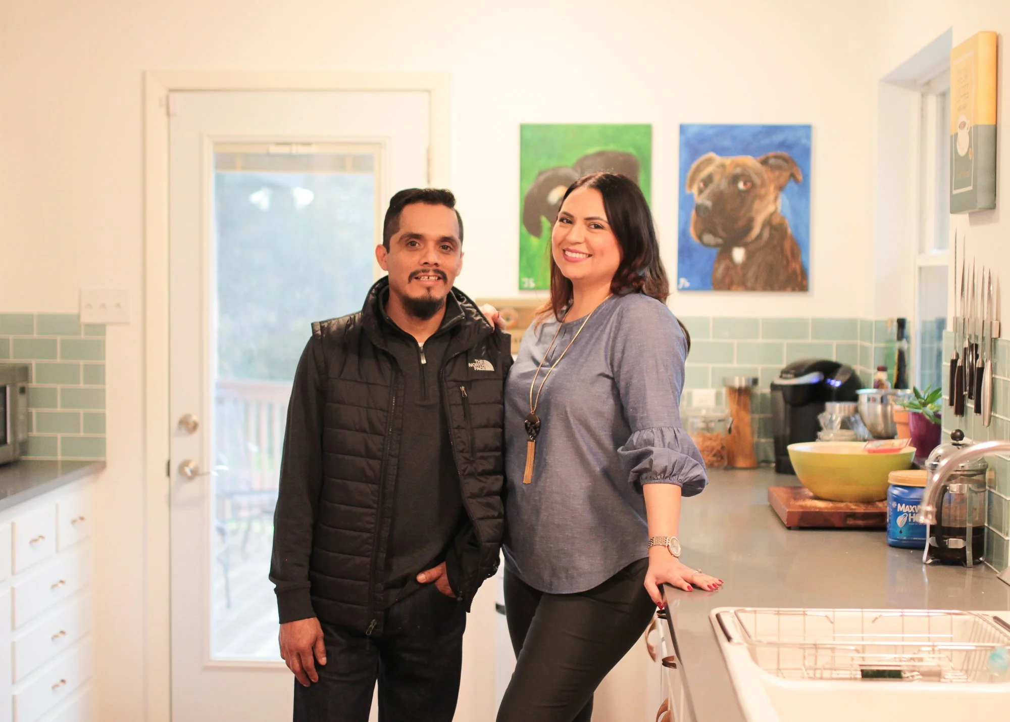 A man and woman standing together in a kitchen, smiling at the camera. The woman has her hand on the counter. The kitchen has green tile backsplash, artwork of dogs on the wall, and various kitchen items on the counter.