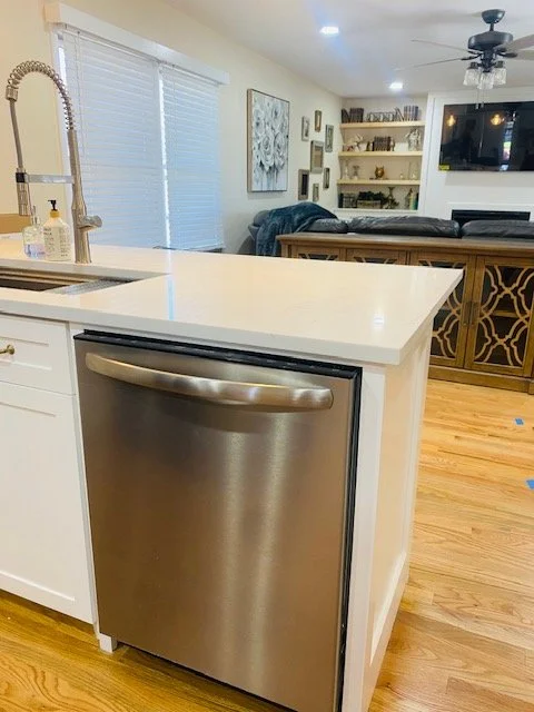 Kitchen with white counter, stainless steel dishwasher, and wooden floor. Living area with shelves and decor in the background.