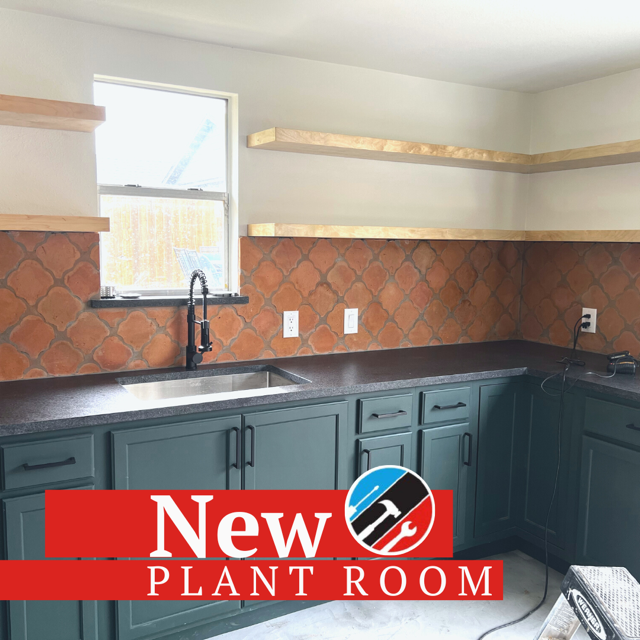 Renovated kitchen with blue cabinets, dark countertop, terracotta tile backsplash, open wooden shelves, and a window above the sink.