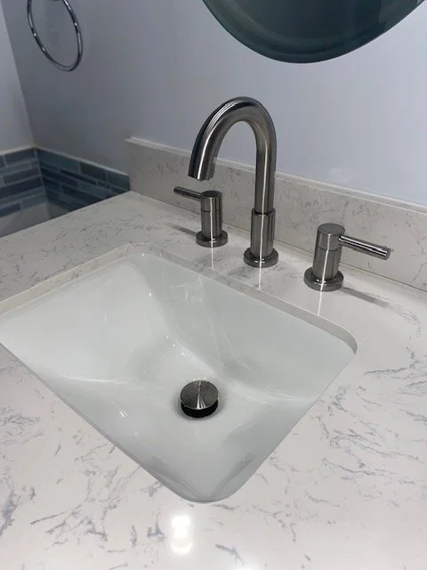 Bathroom sink with a marble countertop, modern silver faucet, and two handles, with a round mirror above and a dark tiled wall in the background.