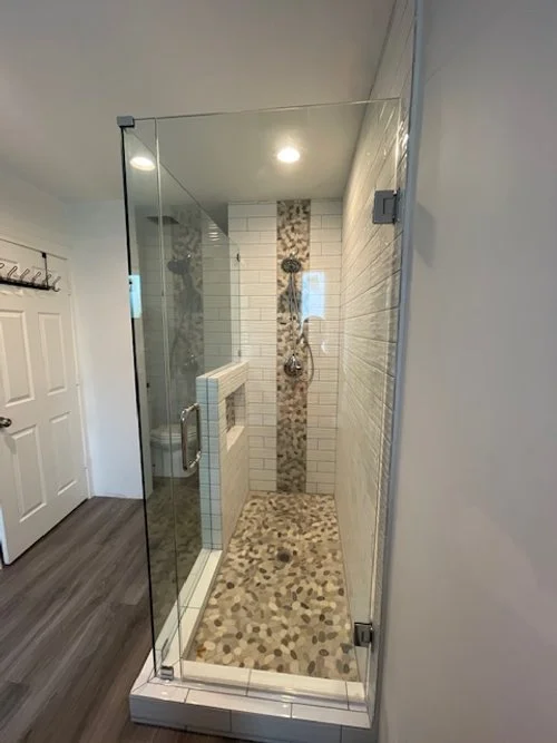 A glass-enclosed walk-in shower with pebble tile floor, white tiled walls, and a decorative vertical strip of patterned tiles in the shower.