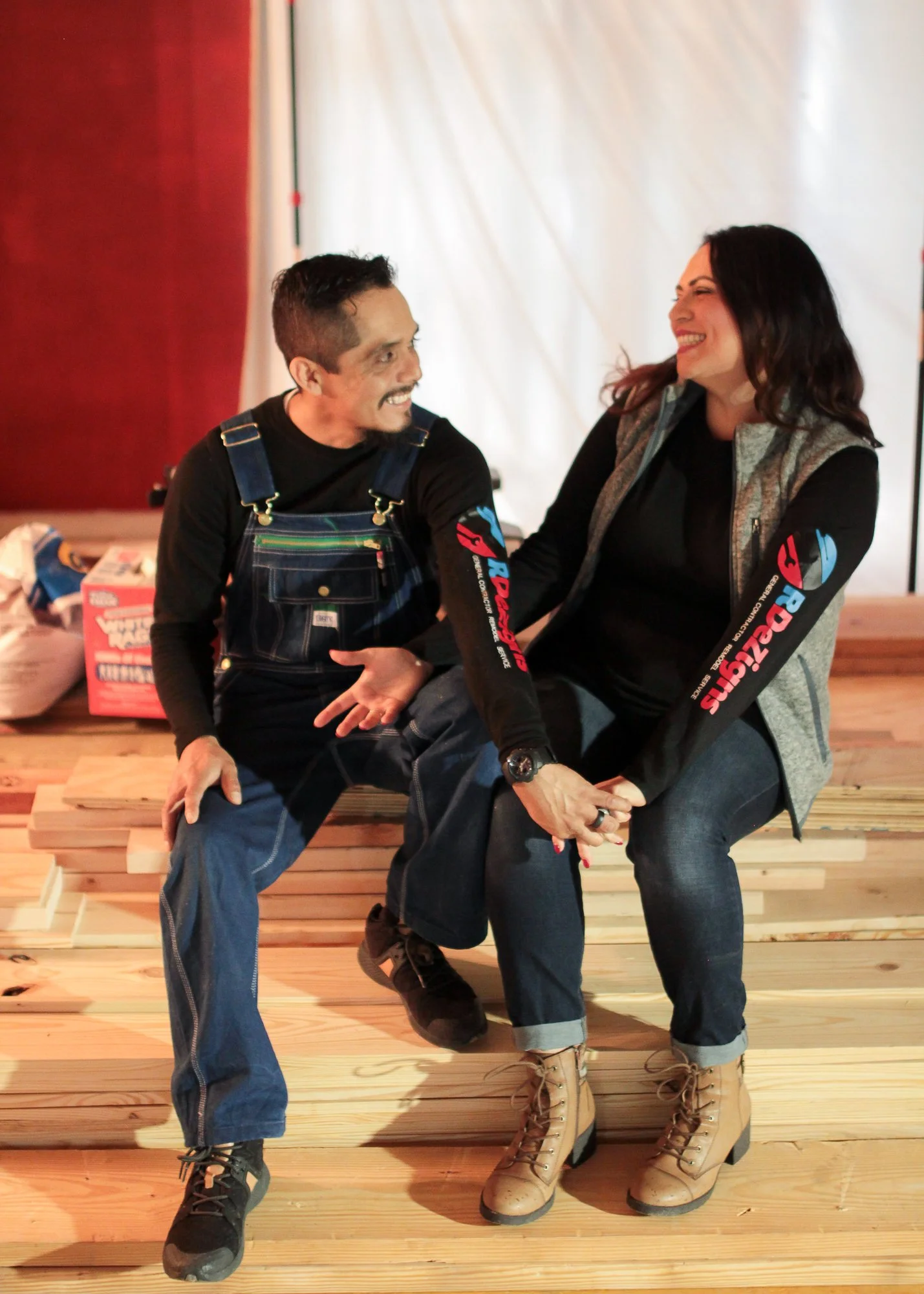 Two people sitting on a wooden bench, smiling and holding hands, with a woman on the right and a man on the left, both wearing casual clothing with matching sleeves.