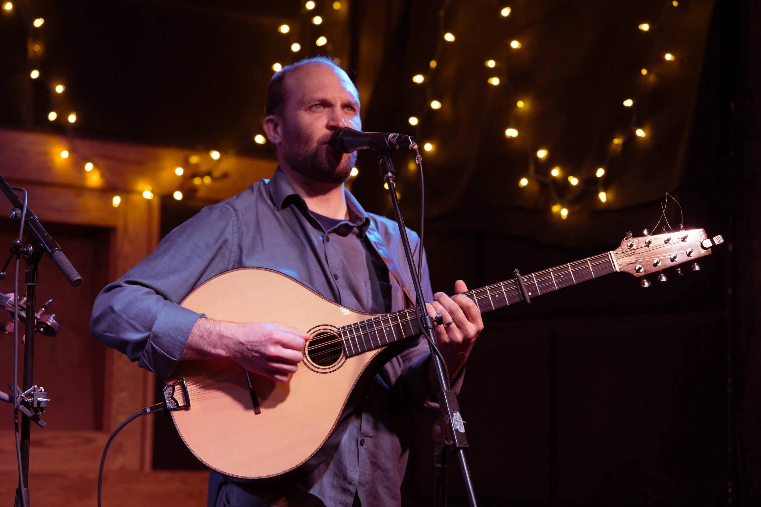 Man performing with guitar and microphone on stage, decorated with string lights.