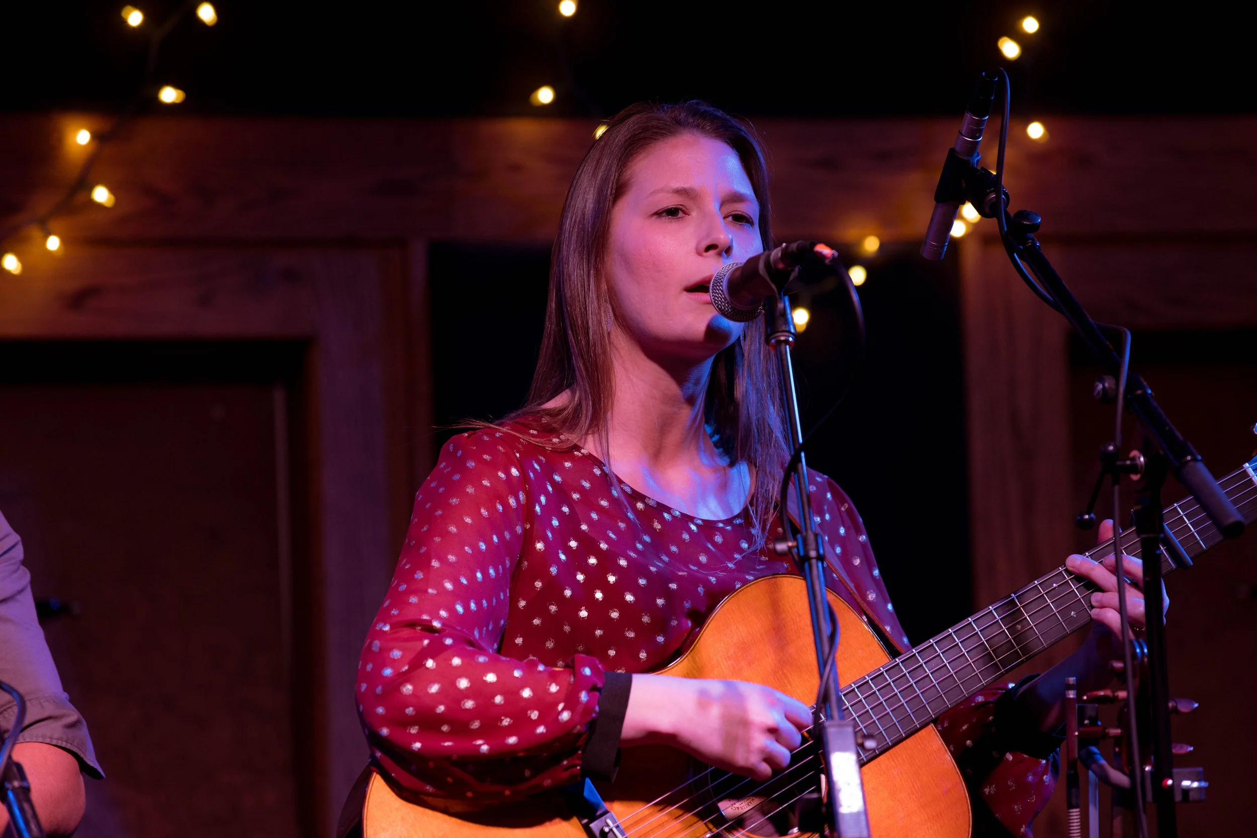 A woman with long brown hair singing into a microphone while playing an acoustic guitar during a live performance, with string lights in the background.