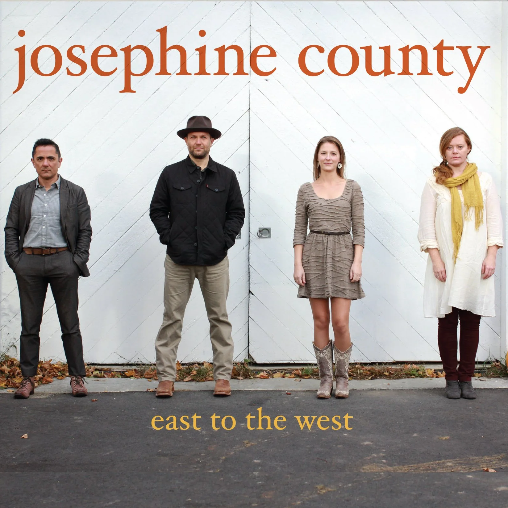 Four people standing in front of a white wall with large text that reads 'josephine county' at the top and 'east to the west' at the bottom. The individuals are evenly spaced, facing the camera with neutral expressions.