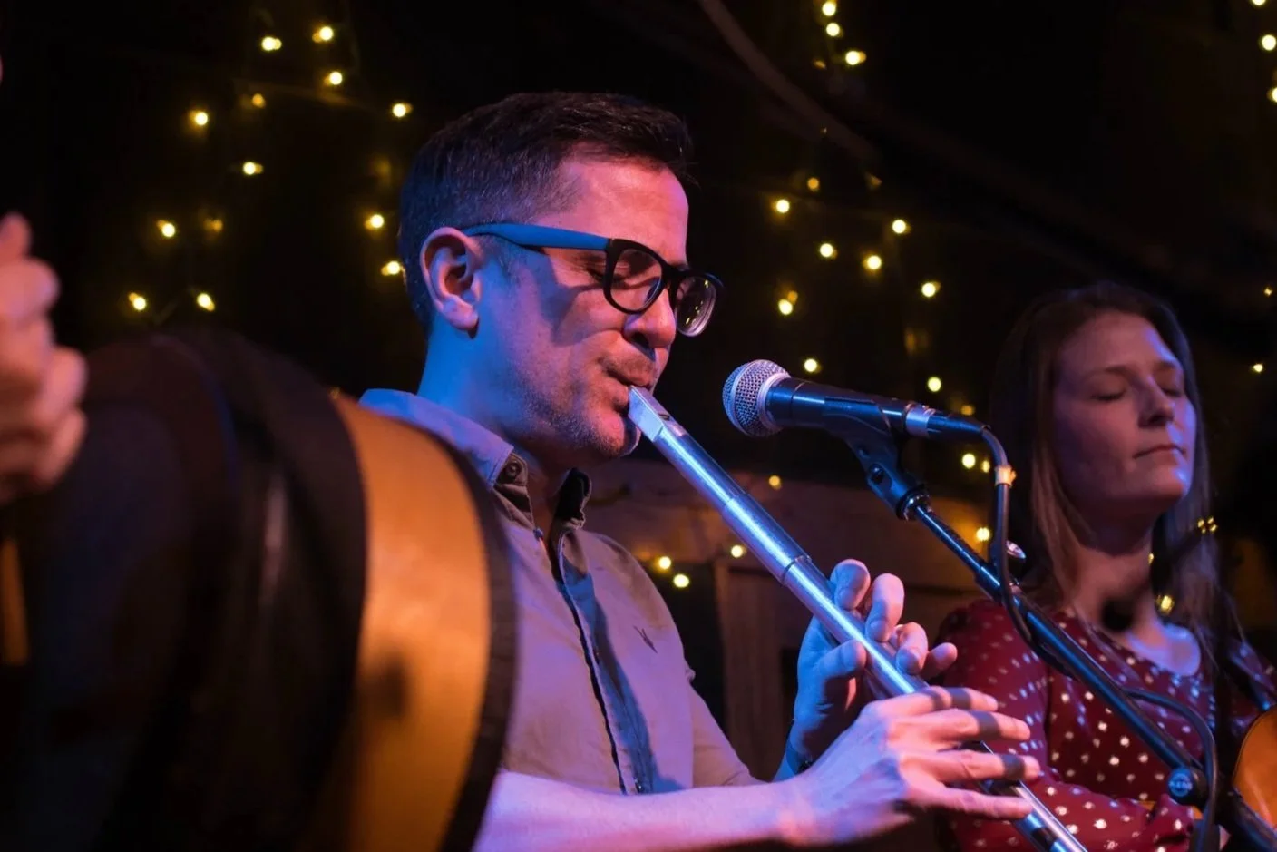 Two musicians performing on stage; one playing a flute and the other with a guitar, with a background of warm string lights.