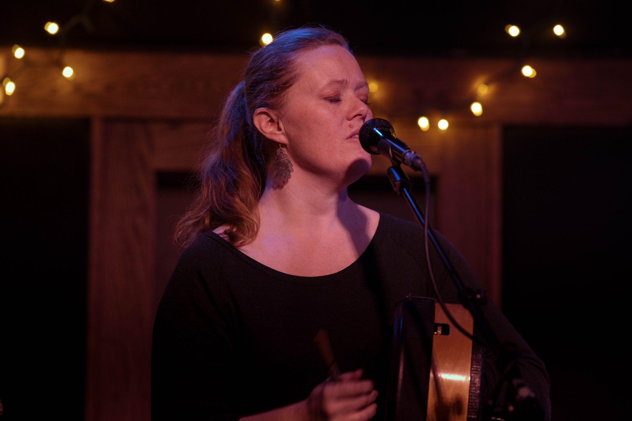 A woman with red hair singing with closed eyes into a microphone on stage, holding a guitar, with string lights in the background.