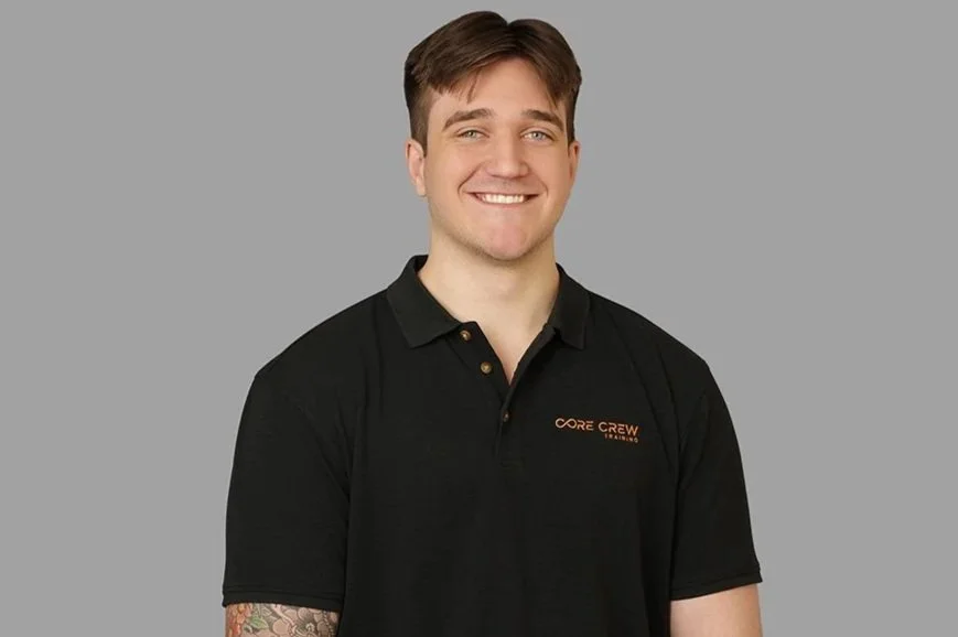 Young man with short brown hair wearing a black polo shirt with orange text, smiling against a gray background.