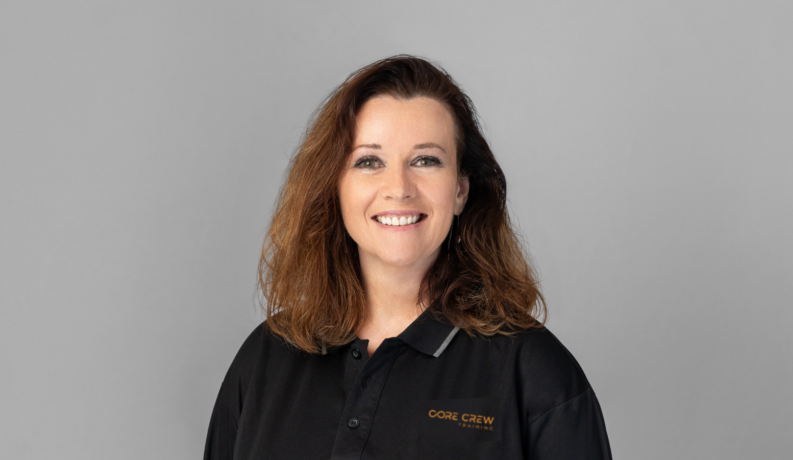 A woman wearing a black polo shirt with 'Core Crew' logo smiling against a gray background.