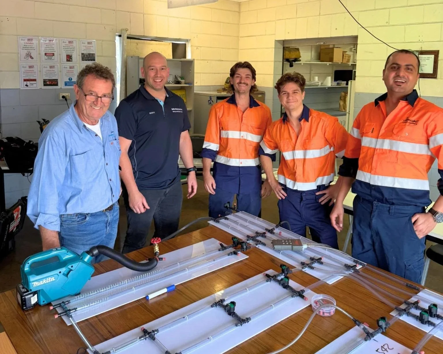 Group of five men standing around a table with various equipment, including a Makita tool, in a workshop setting. Three men are wearing orange safety shirts, one in a blue shirt, and one in a dark polo.