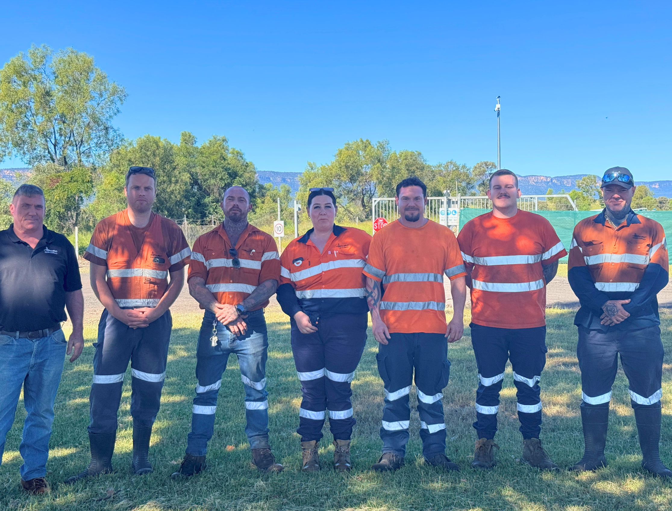 A group of seven people standing outdoors on grass, wearing orange and black uniforms with reflective stripes, with trees, a fence, and mountains in the background on a clear, sunny day.