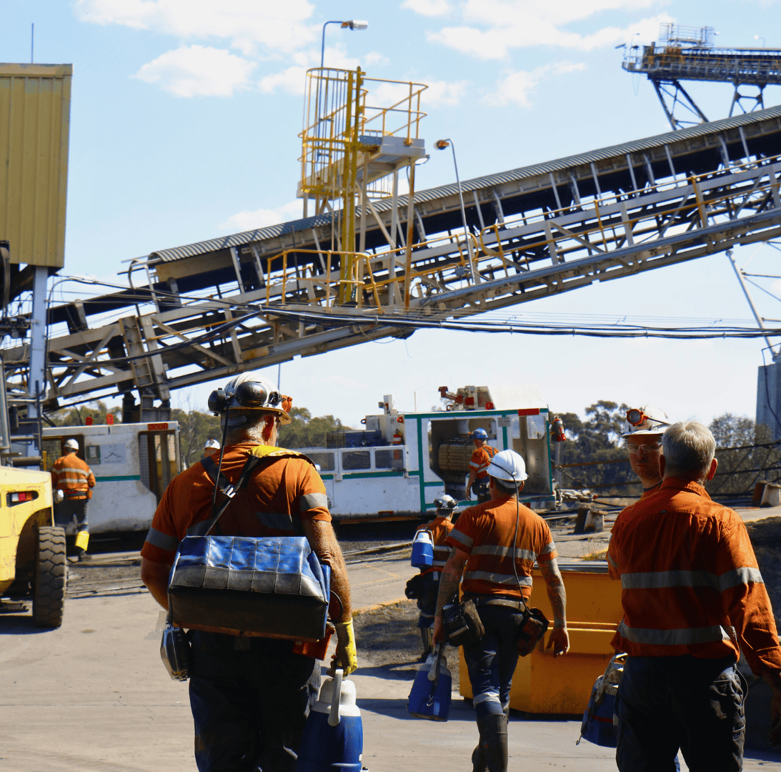 Construction workers in orange safety uniforms working on industrial site with machinery and conveyor belt system under a partly cloudy sky.