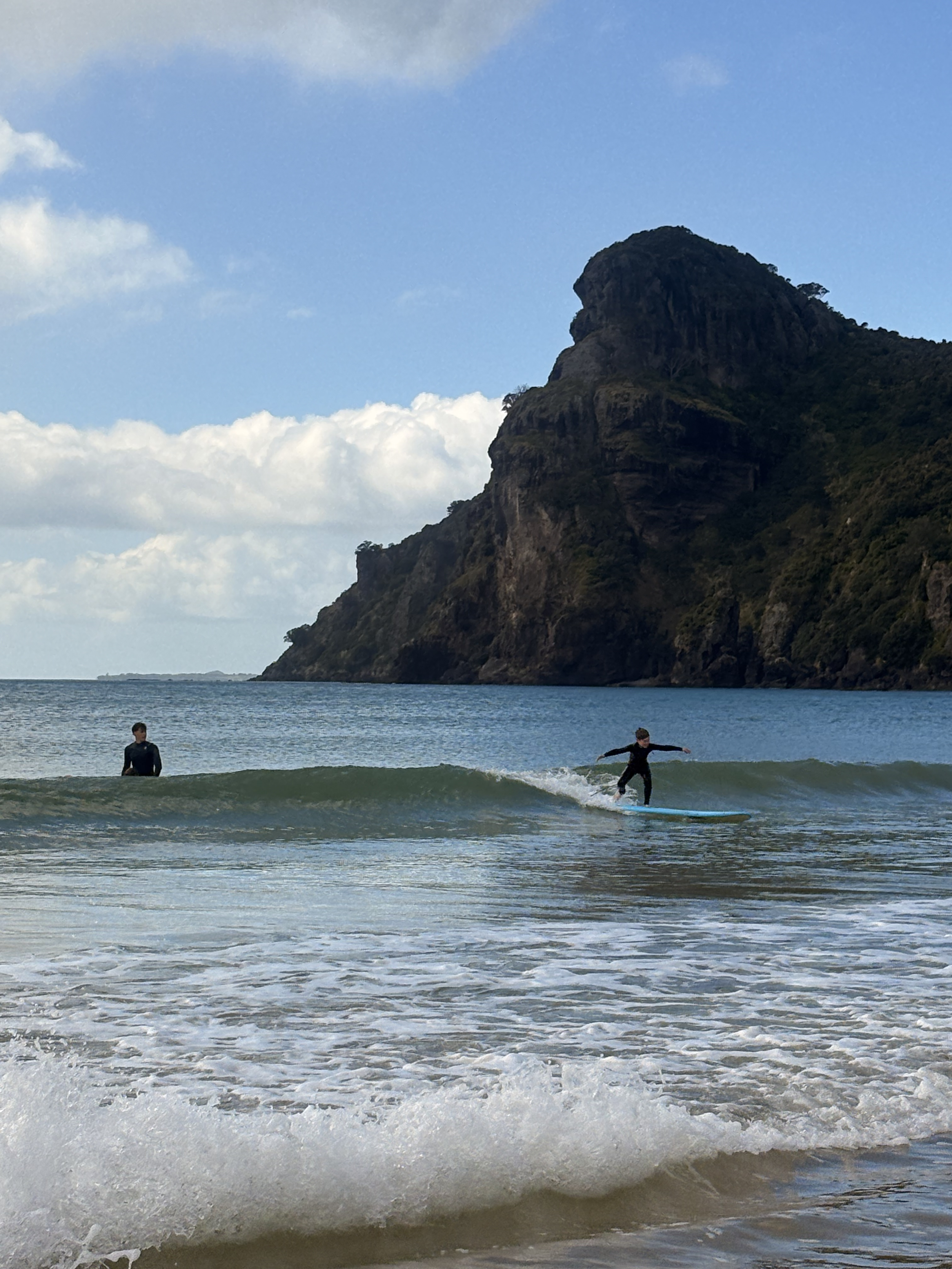 A young surfer riding a wave at Taupō Bay with the headland behind