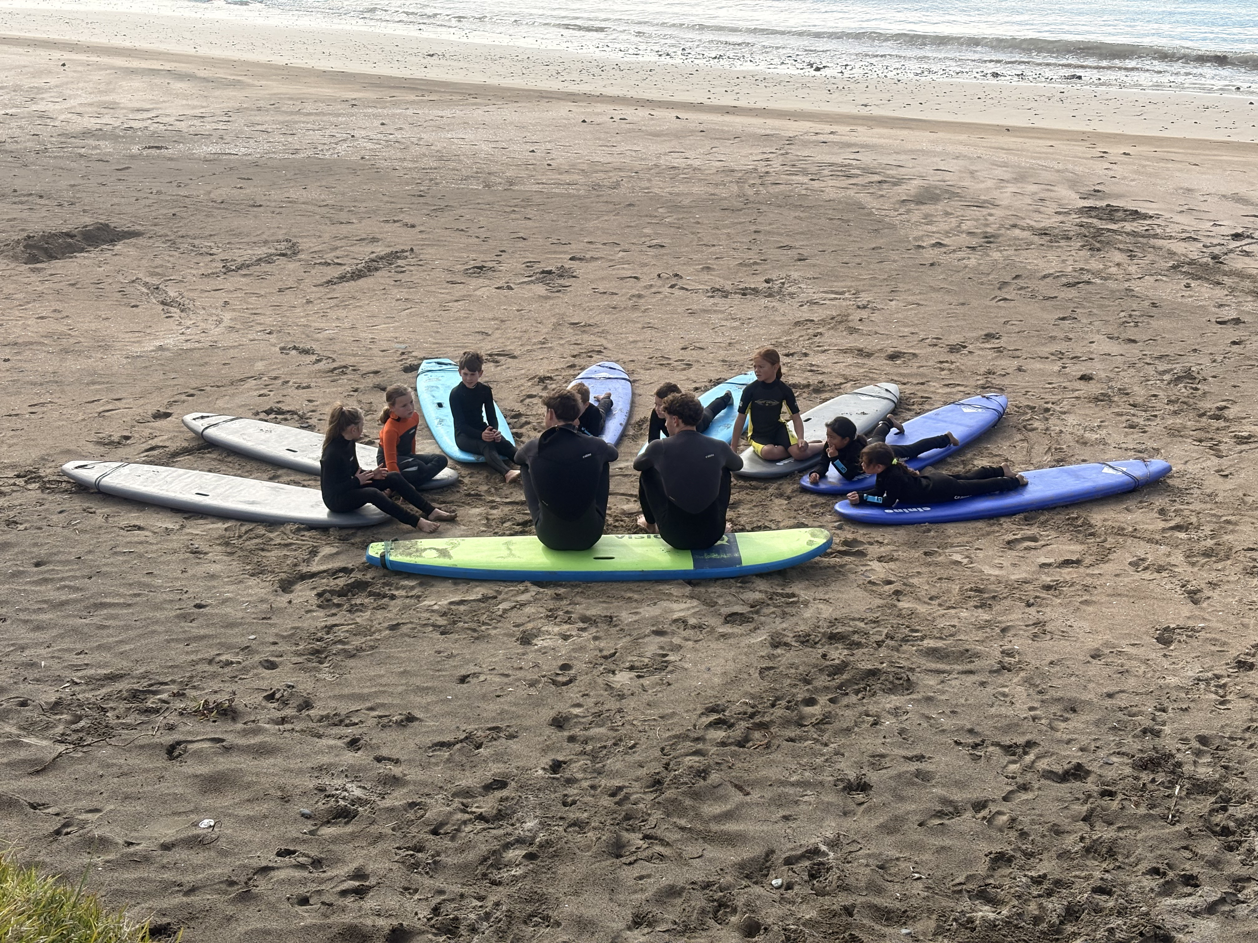 A group of children and adults sitting and lying on surfboards on a sandy beach, with ocean waves in the background.