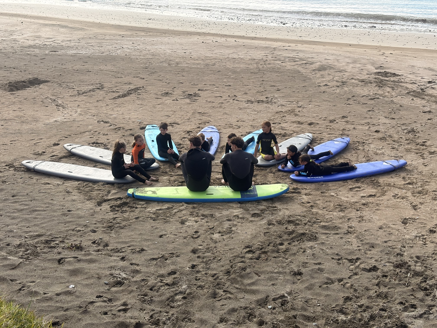 Kids in wetsuits sitting in a circle on their surfboards on the sand at Taupō Bay, listening to their surf coach