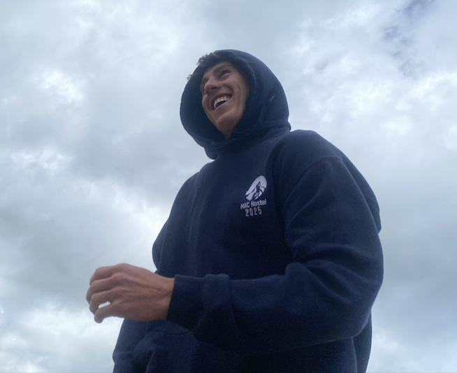 Young man smiling in a hoodie outdoors against cloudy sky.