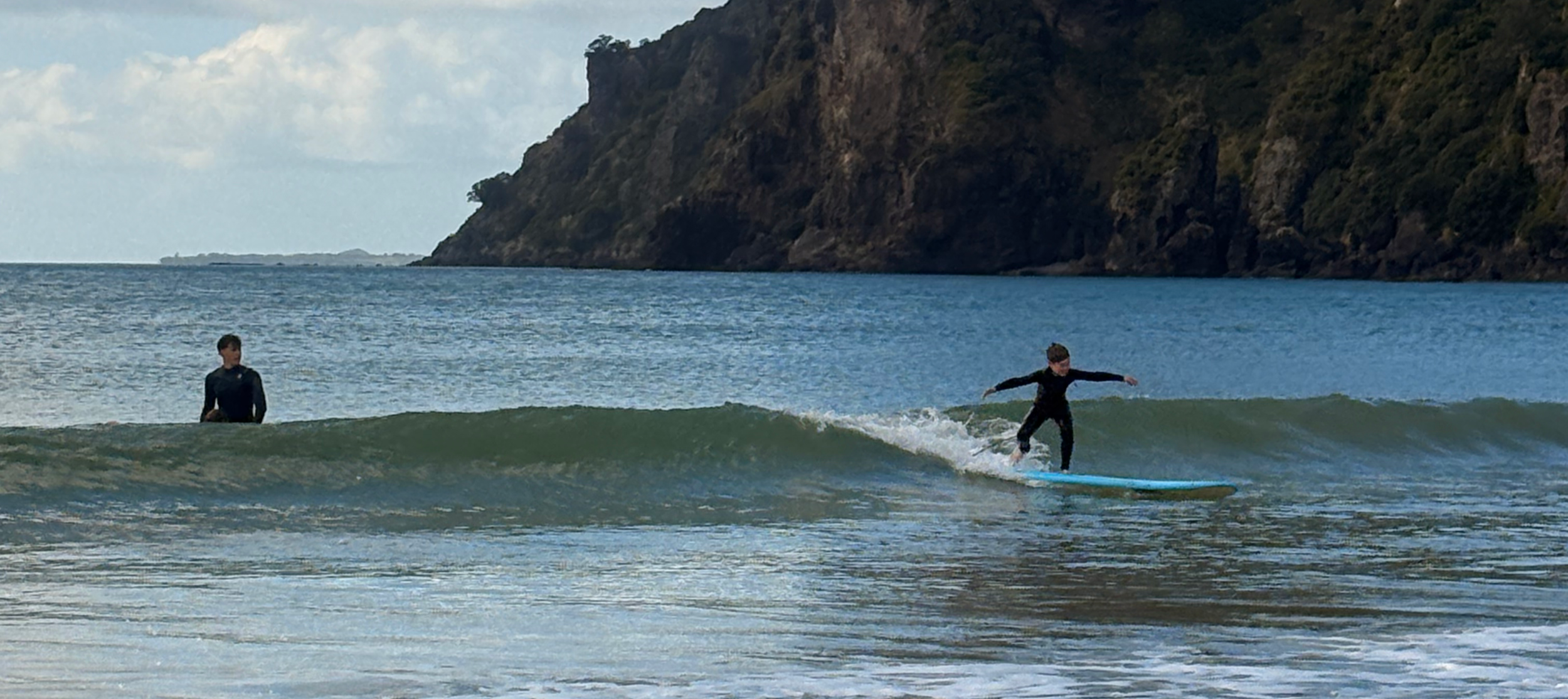 Two children in wetsuits, one standing in the water and the other riding a wave on a surfboard at a beach with a rocky cliff in the background.