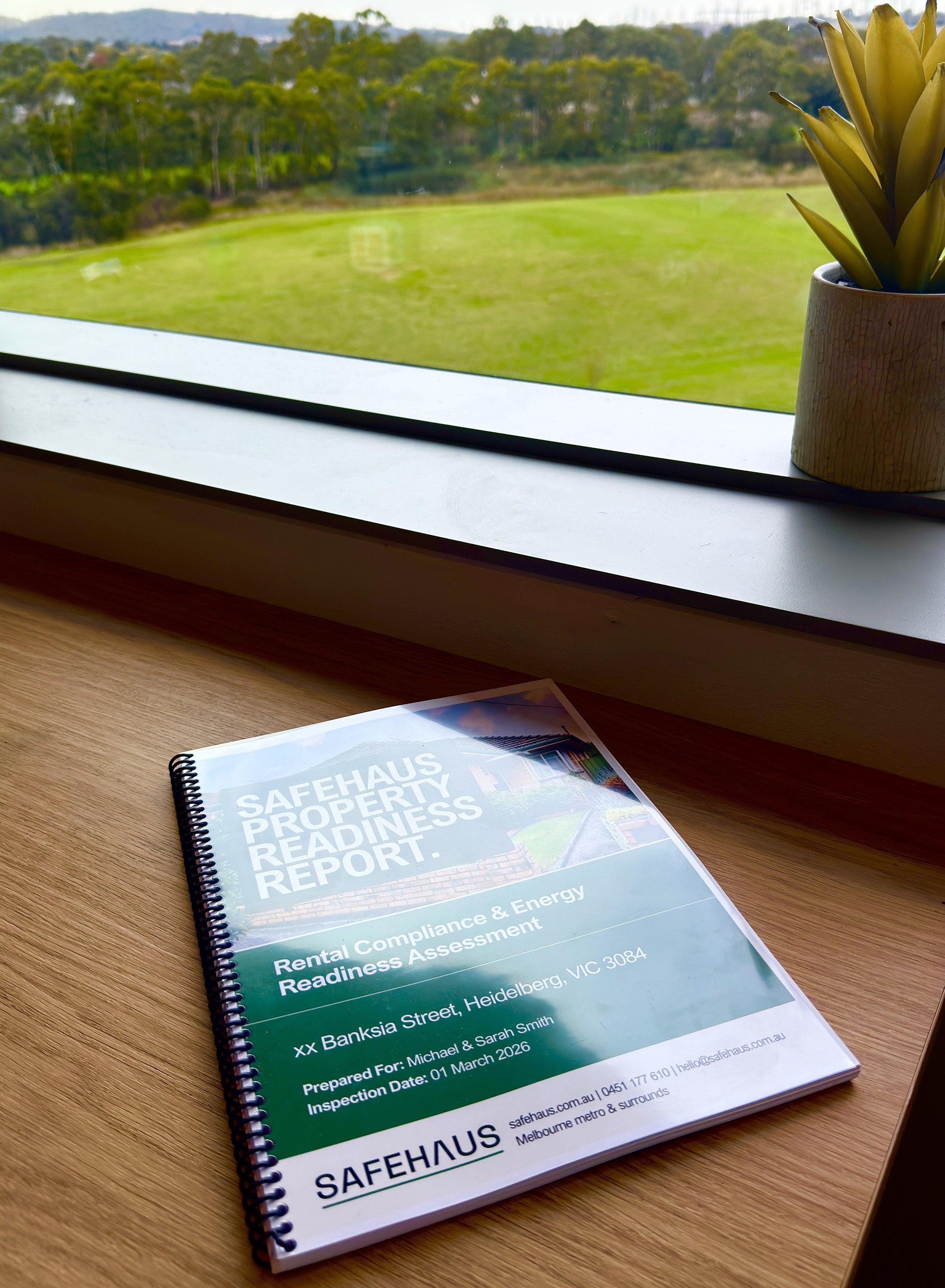 A spiral-bound report titled "Safehaus Property Readiness Report" resting on a wooden desk, with a window in the background showing a green grassy landscape and trees.