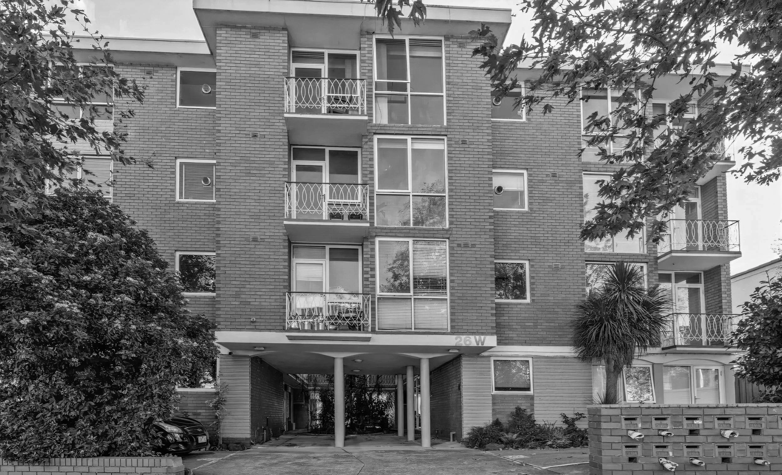 Front view of a multi-story apartment building with balconies, surrounded by trees and bushes, black and white photograph.