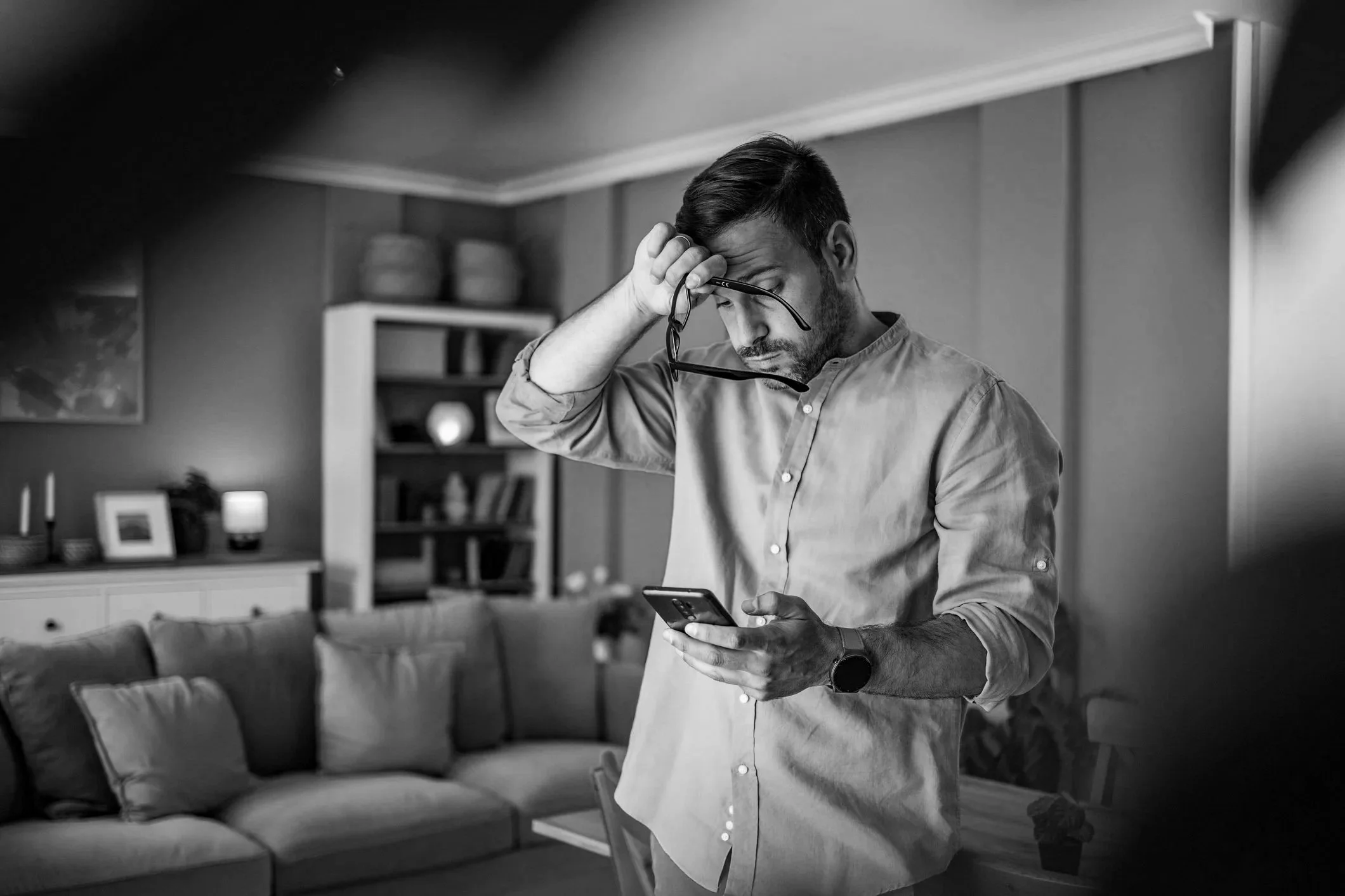 A man standing in a living room looking at his phone while holding his glasses, with a distressed expression on his face.
