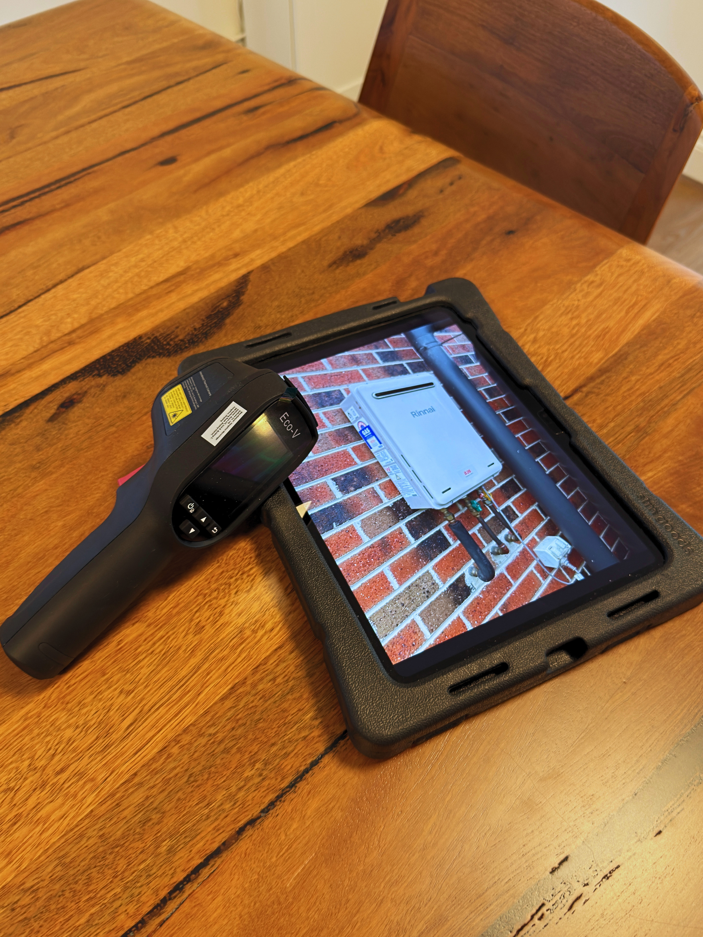 Tablet showing a brick wall with an outdoor scene, a handheld device labeled 'ECO V', and a black case or tray on a wooden table.