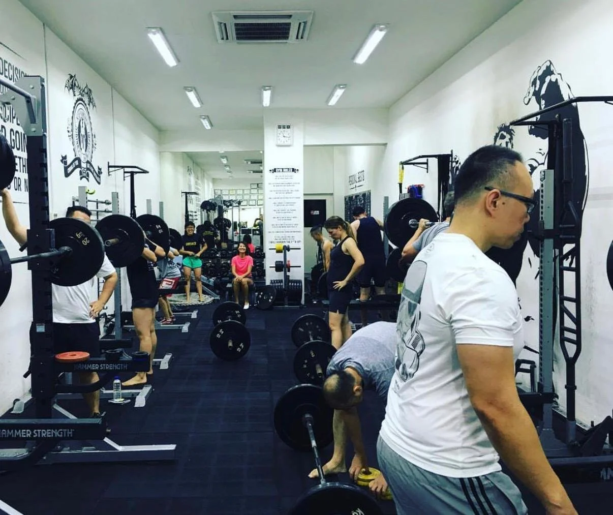 People working out in a gym with weightlifting equipment, including barbells and weight plates, among white walls with gym-themed art and motivational quotes.