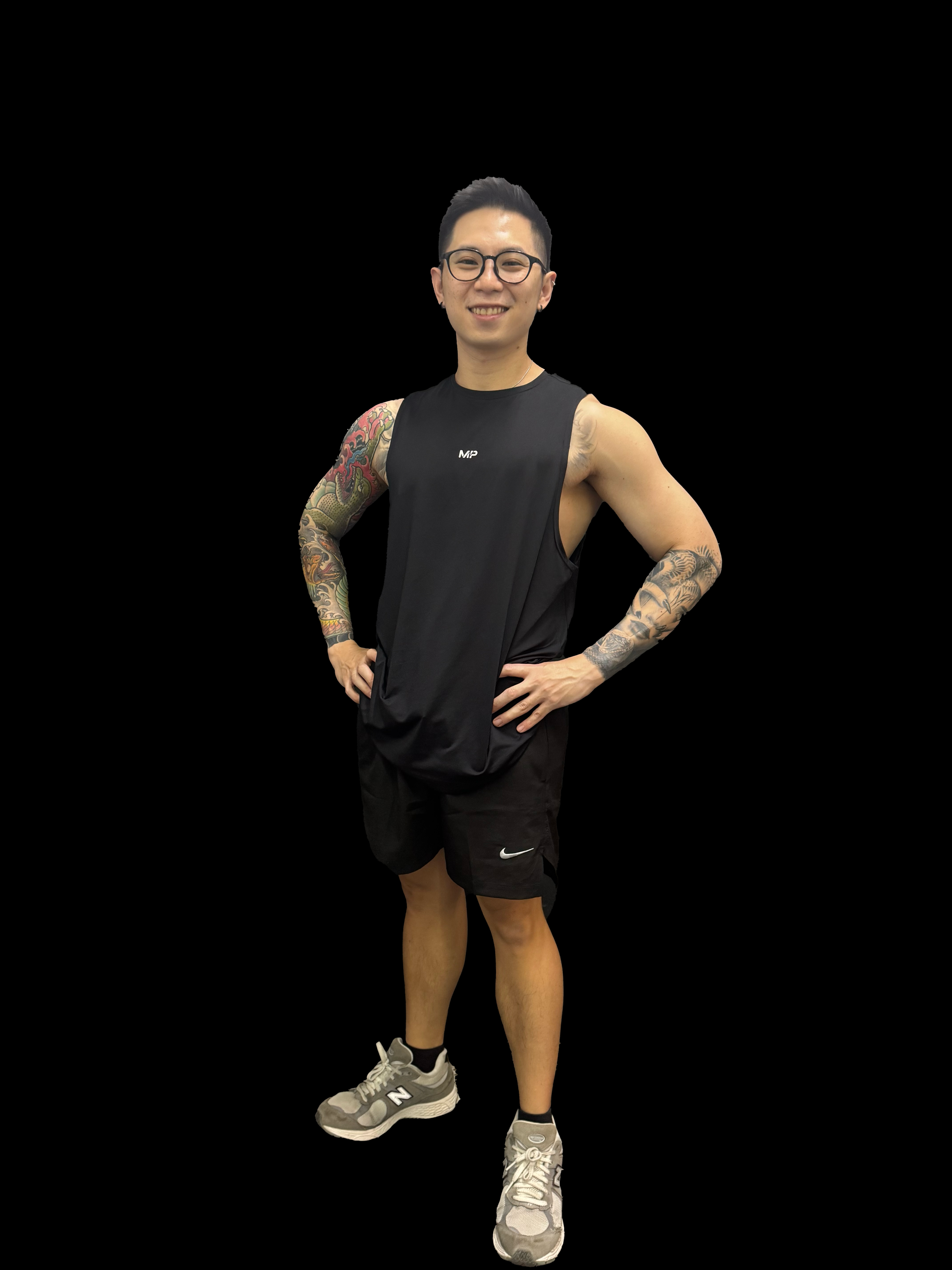A young man with tattoos on both arms, wearing glasses, a black sleeveless athletic shirt, black shorts, and gray sneakers, standing with hands on hips and smiling.