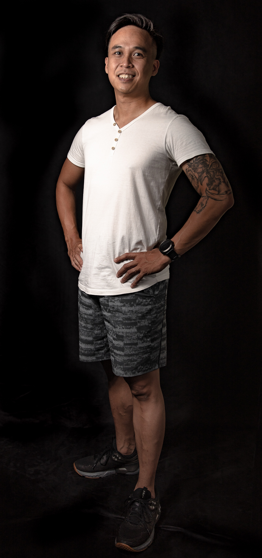 A man standing against a dark background, wearing a white V-neck T-shirt, patterned shorts, and running shoes, with one hand on his hip and smiling.