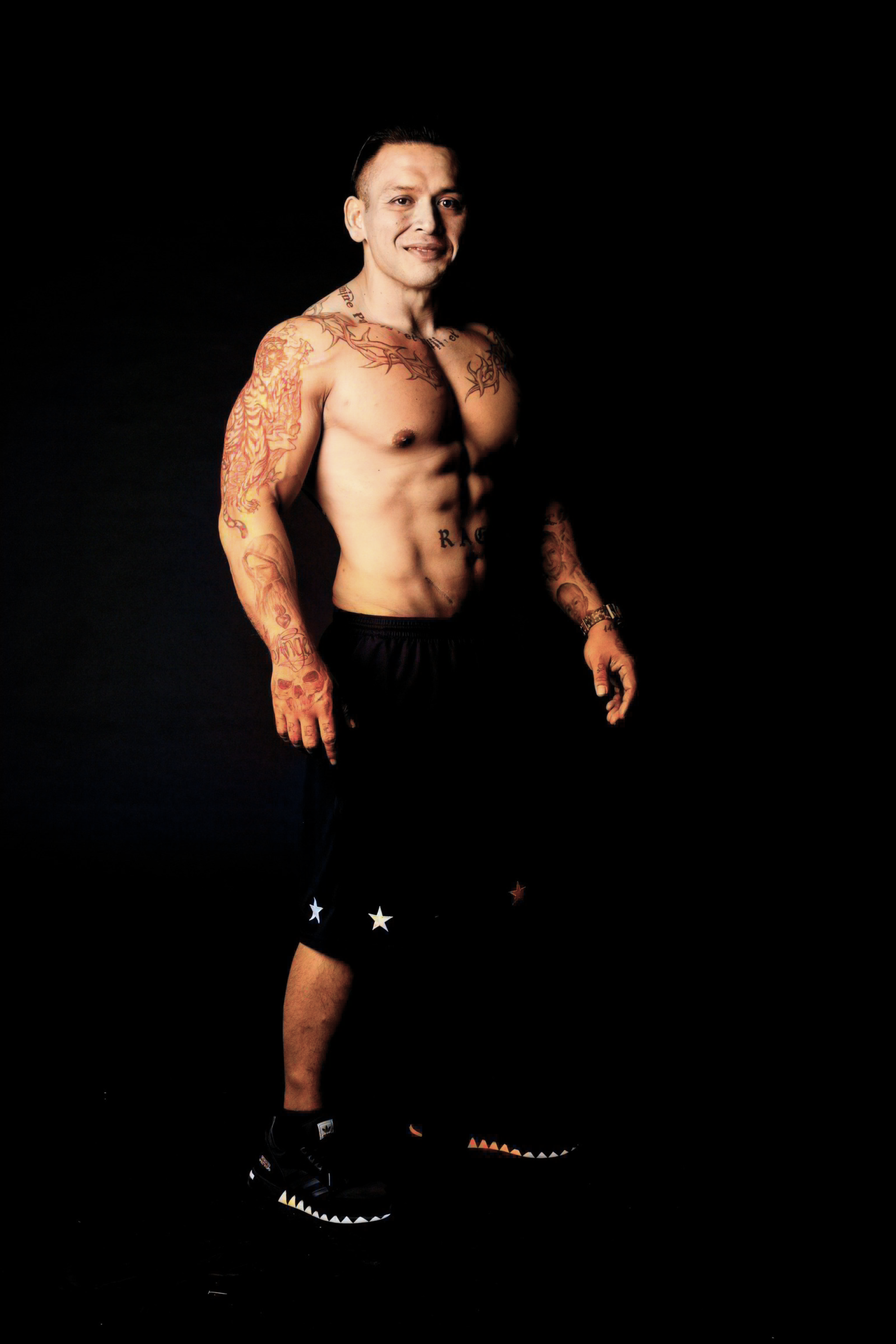 A shirtless man with tattoos on arms and chest, wearing black shorts with star patterns and black sneakers, standing against a black background.