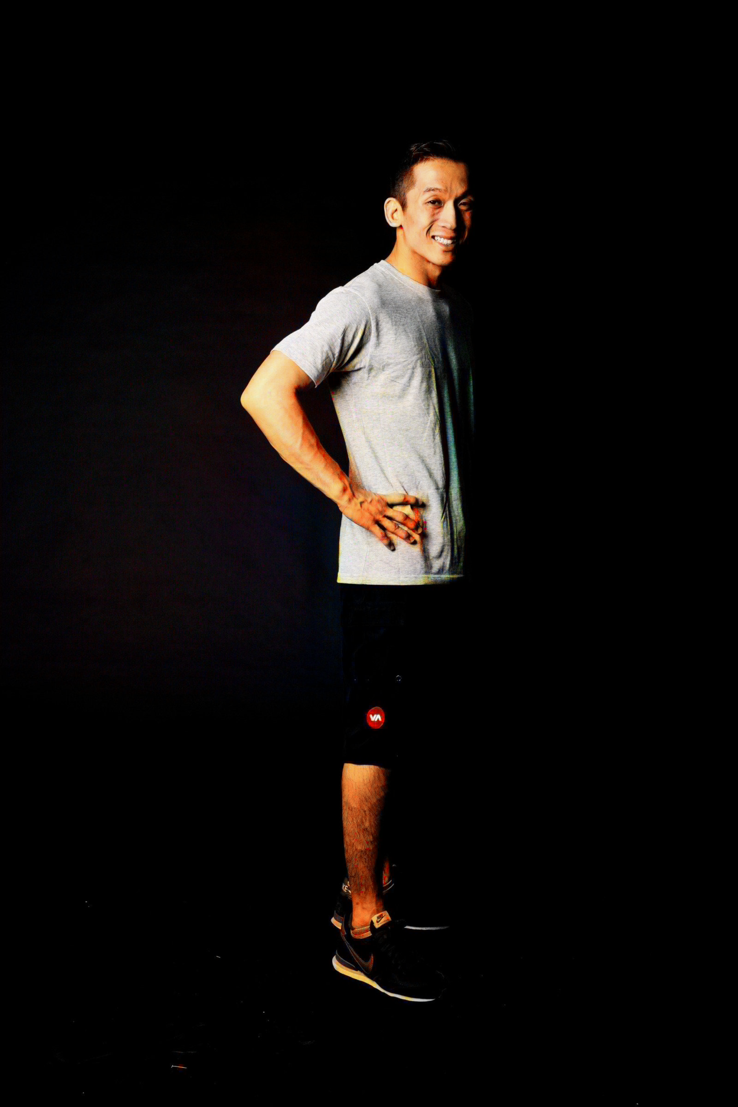 A man standing against a black background, smiling, wearing a gray t-shirt, black shorts with a red logo, and black sneakers.