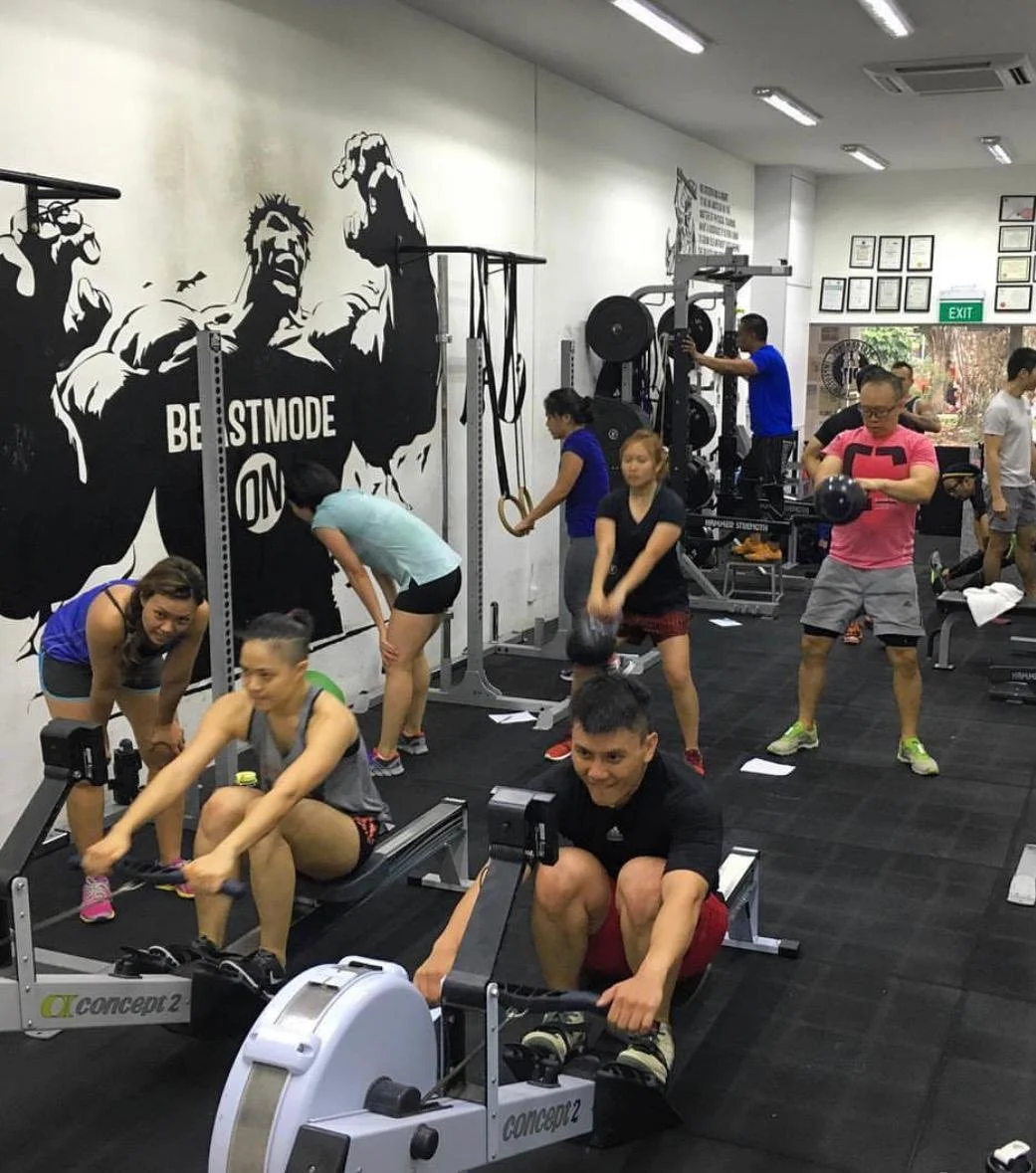 People working out in a gym, including a woman on rowing machine, a woman with gymnastic rings, a man lifting a kettlebell, and others exercising with various equipment.