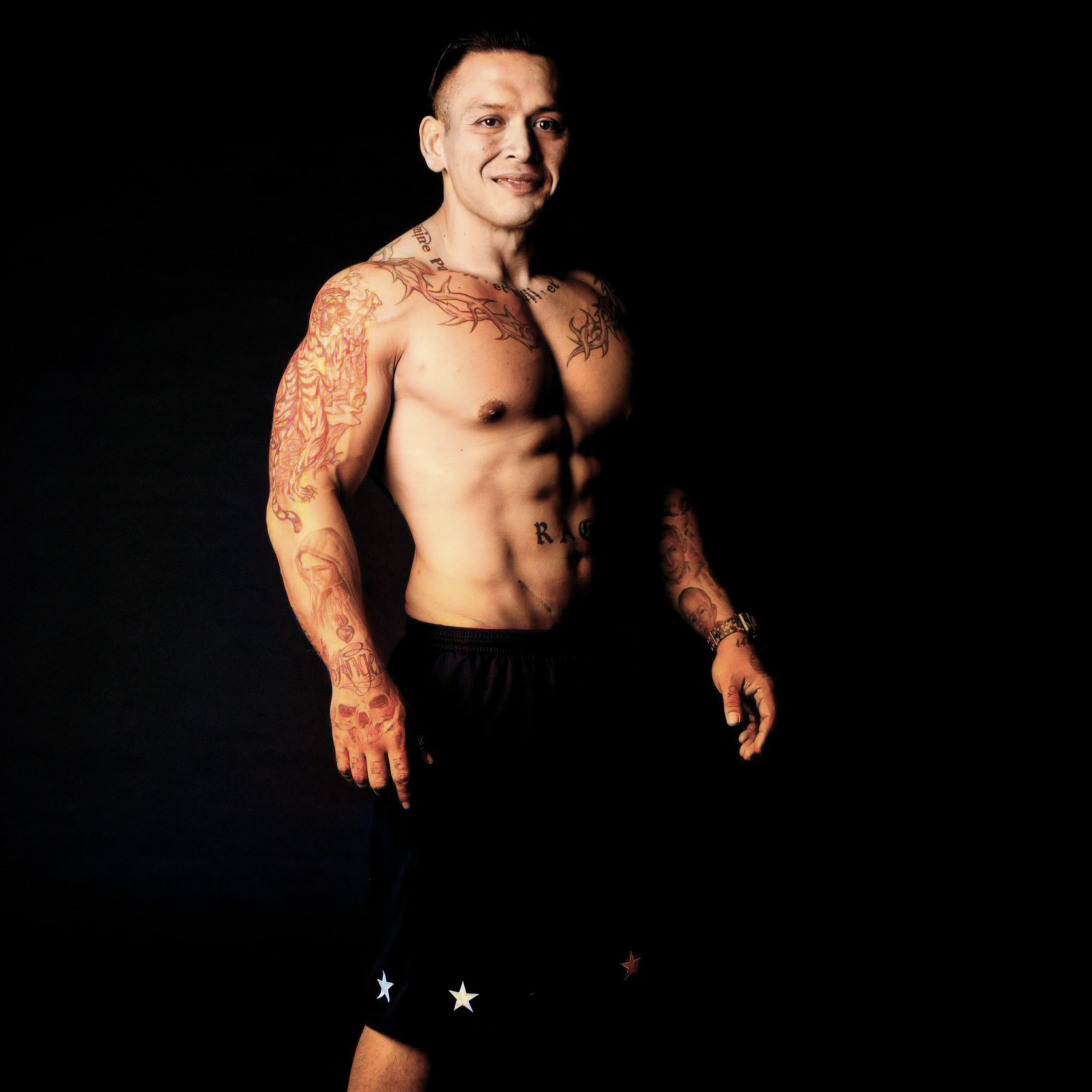 A shirtless man with tattoos standing against a black background, smiling at the camera.
