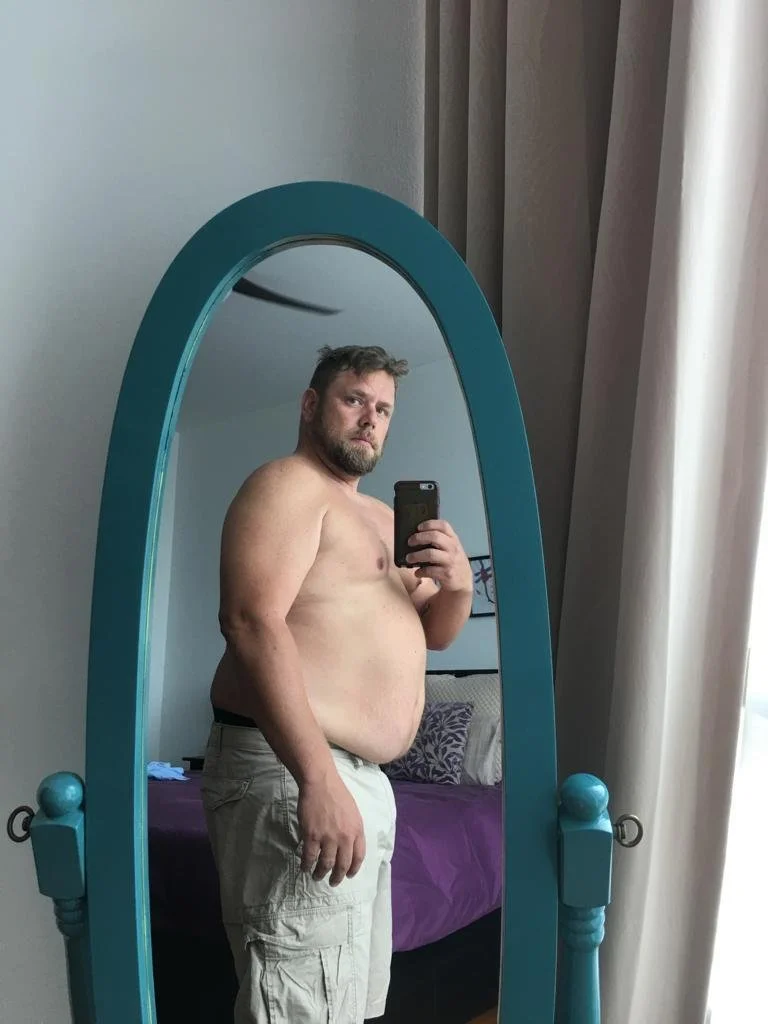 A shirtless man with a beard and khaki shorts taking a mirror selfie in a bedroom, with a bed, purple blankets, and gray curtains in the background.