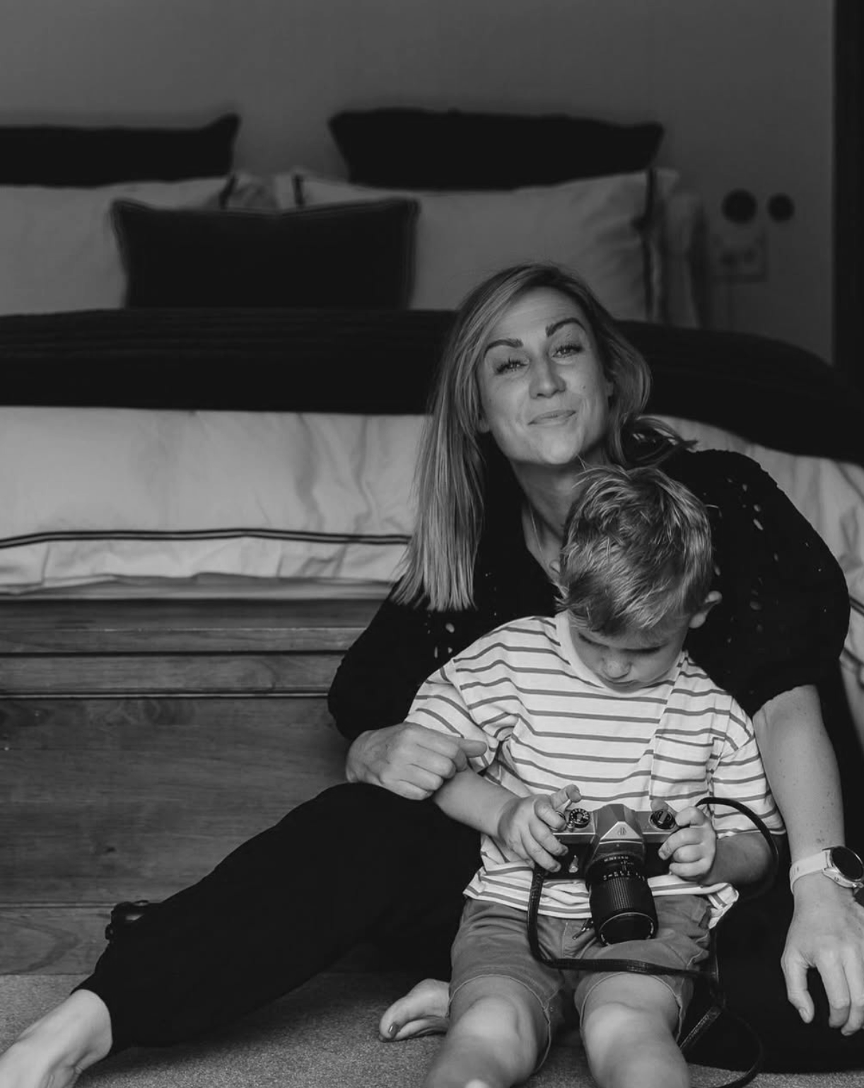 A woman and a young boy sitting on the floor in front of a bed, the boy is looking at a camera he is holding while the woman smiles at the camera.