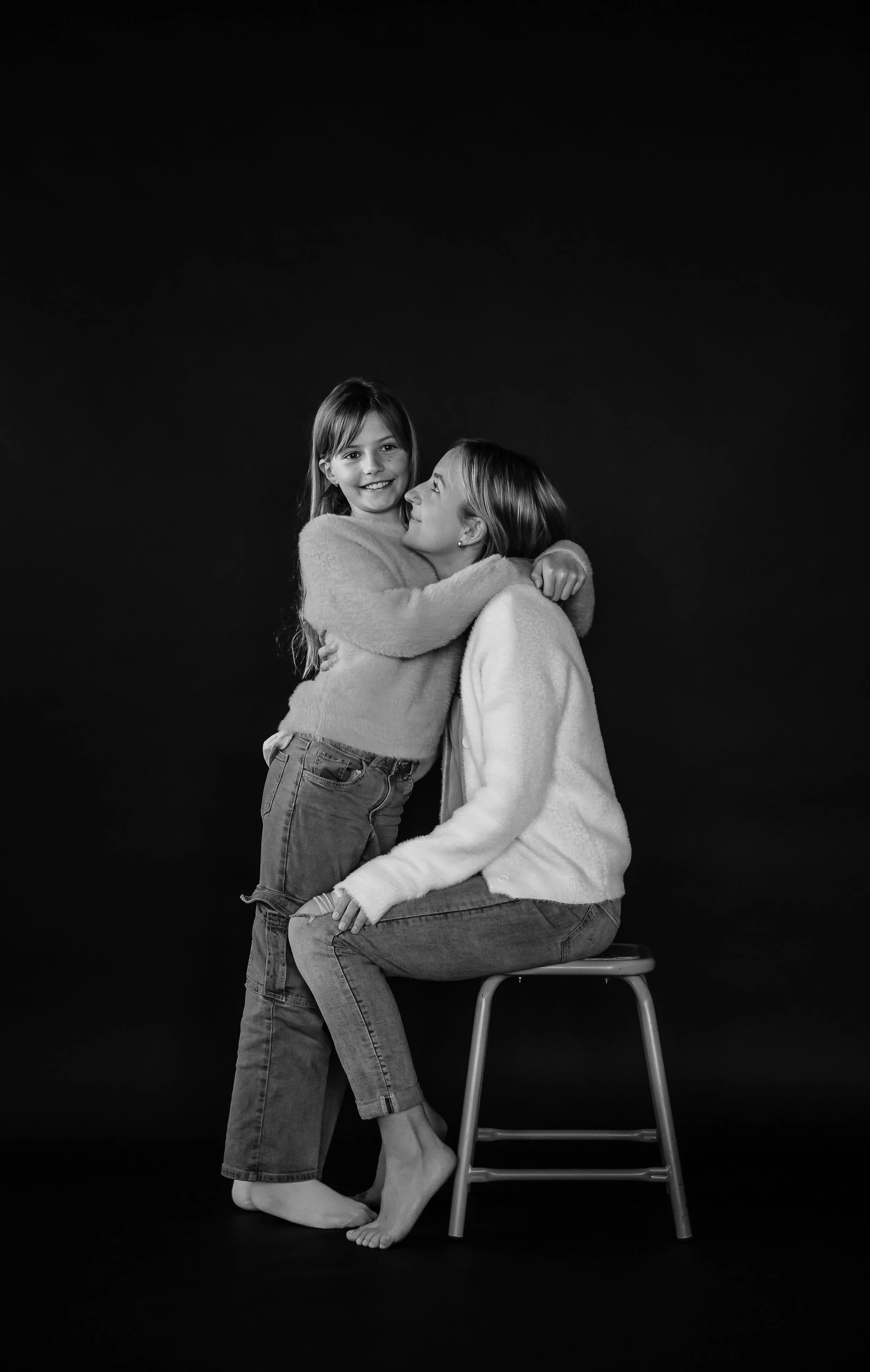 Black and white photograph of a woman sitting on a stool and a young girl hugging her, both smiling, against a dark background.