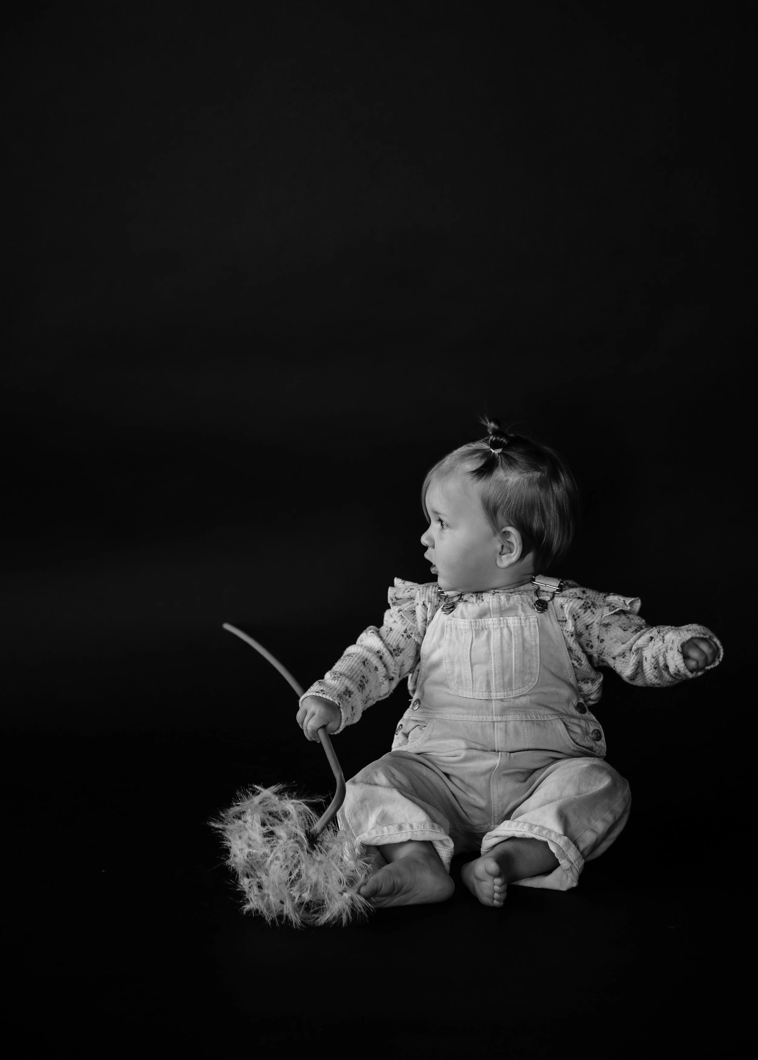 A young child sitting on the floor holding a feather duster, looking to the side, against a dark background.