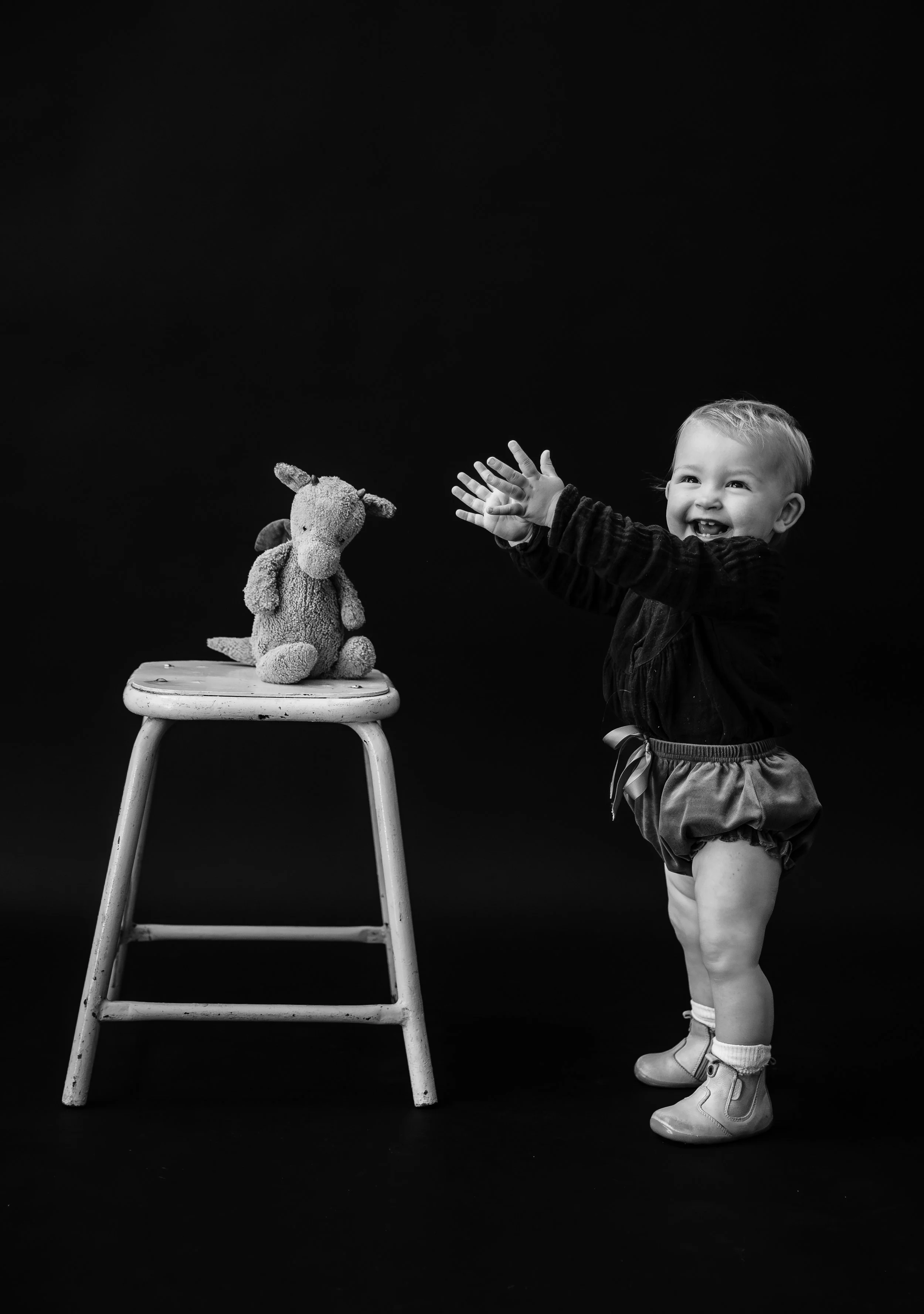 A smiling young child in shorts and boots reaching towards a plush toy on a small stool against a black background.